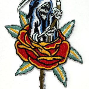 以下が含まれることがあります： 鎌を持った死神の姿が描かれた刺繍パッチ。赤と黄緑色のアクセントが施された赤いバラから現れています。死神は白黒で、青いフードをかぶっています。衣類やアクセサリー用です。