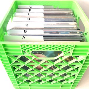 May include: A bright green plastic crate filled with CDs, organized with white divider tabs labeled A through G. The CDs are in their cases, and the crate has a diamond-patterned design.
