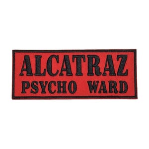 May include: A rectangular patch with the words "ALCATRAZ PSYCHO WARD" in black, embroidered on a red background. The patch has a black border and is likely for clothing or accessories.