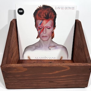 May include: A dark brown wooden record holder displaying a vinyl record. The record features David Bowie's "Aladdin Sane" album cover. The cover art shows David Bowie with red hair and a lightning bolt across his face. The holder is made of wood.
