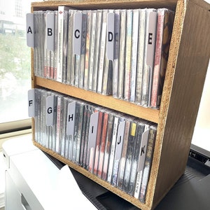 May include: A wooden CD storage rack with two shelves, each holding multiple CD cases. The cases are labeled with white tabs marked A to K. The CD cases are in clear plastic cases. The text on the tabs is visible.