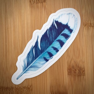 May include: A blue jay feather sticker with a white background and a blue and white design. The sticker has the text "Blue Jay by Misty Stone" in the upper right corner.
