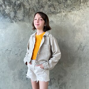 May include: A young person wearing a light beige cropped jacket over a yellow shirt and white denim shorts. The person is standing in front of a textured gray wall.