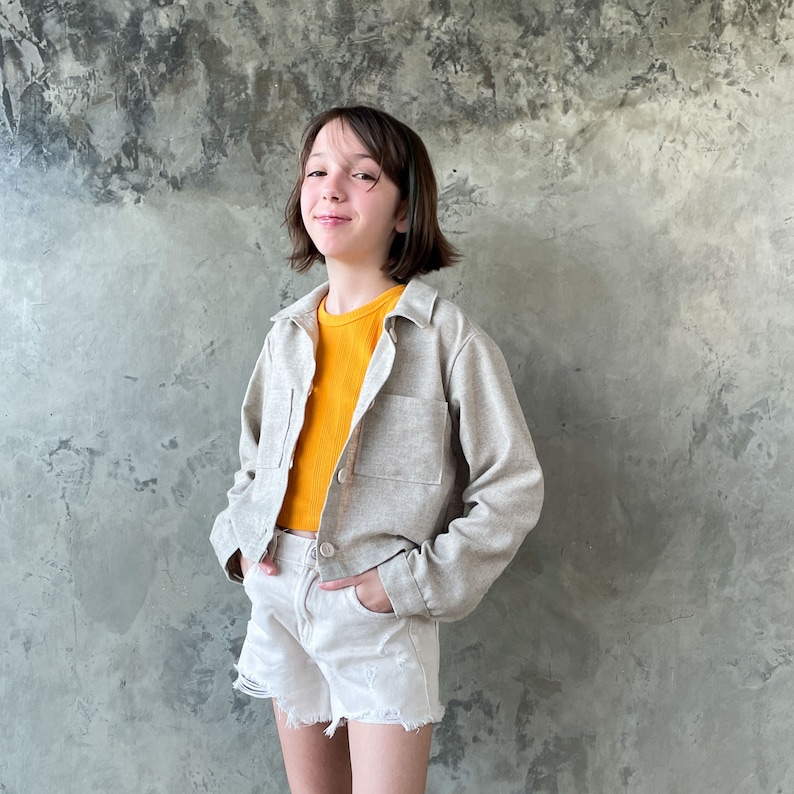 May include: A young person wearing a light beige cropped jacket over a yellow shirt and white denim shorts. The person is standing in front of a textured gray wall.