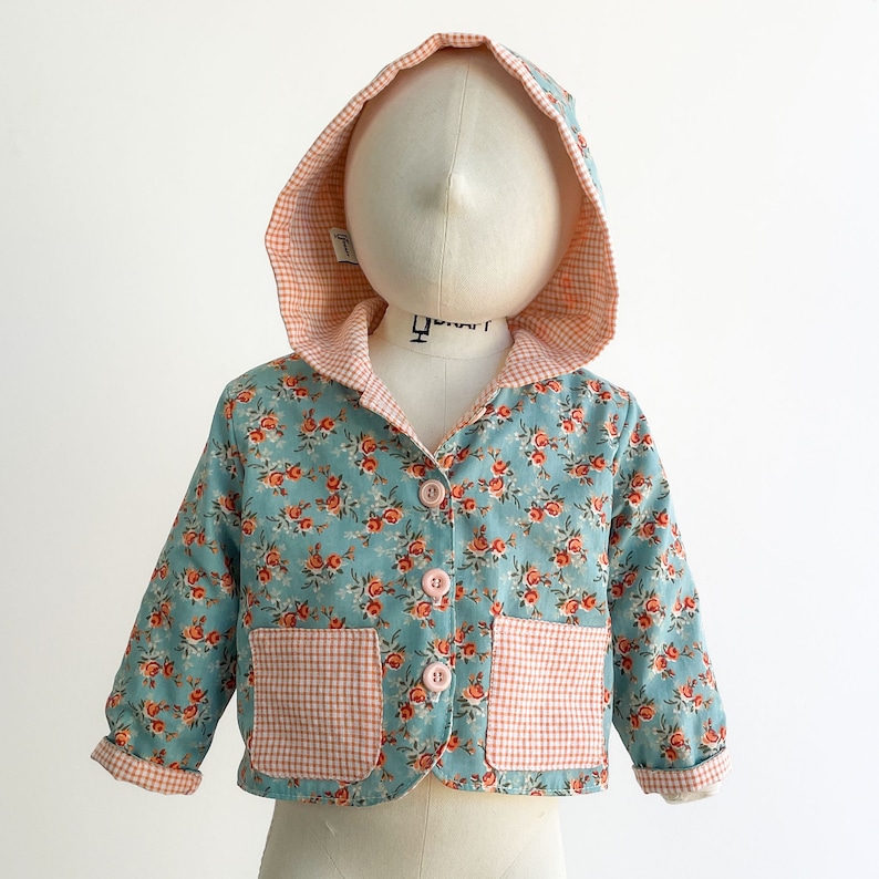 May include: A light blue hooded jacket with a floral print and pink and white gingham pockets. The jacket has a button closure.