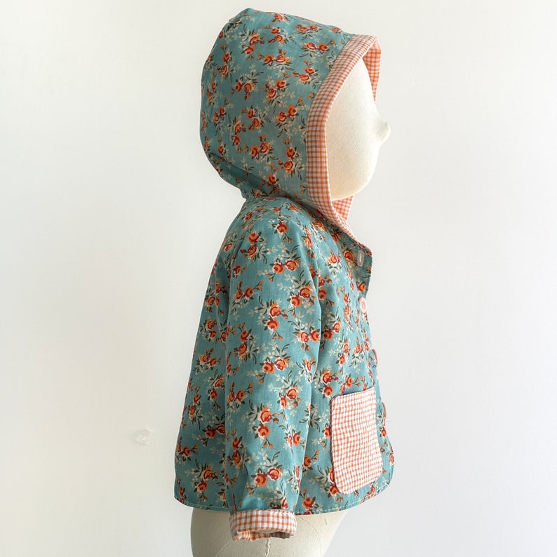 May include: A light blue hooded jacket with a floral print and a red and white gingham pocket. The jacket has a red and white gingham trim around the hood.