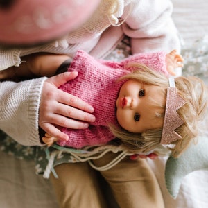 May include: A child holds a doll wearing a pink knitted sweater and a sparkly pink crown. The doll has blonde hair and blue eyes.