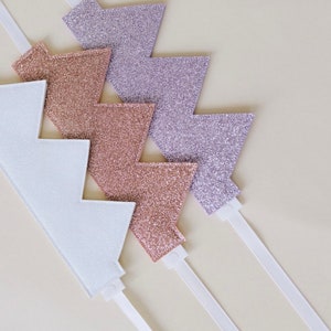 May include: A collection of glittery lightning bolt-shaped hair accessories in white, rose gold, and silver. Each bolt is attached to a thin white headband. The accessories are arranged on a light beige surface.