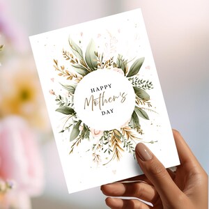 May include: A white Mother's Day greeting card with a floral design. The card features a circular arrangement of green leaves, gold accents, and pink flowers around the text "Happy Mother's Day" in elegant script.