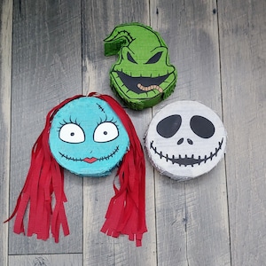 May include: Three colorful pinata decorations featuring characters from the Nightmare Before Christmas. The first pinata is green and shaped like a head with a toothy grin. The second pinata is blue and features a female character with red hair. The third pinata is white and features a skull with a toothy grin.