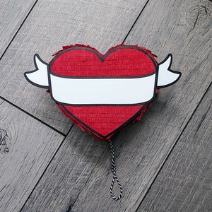 May include: A red heart-shaped piñata with a white banner across the center and white wing-like accents. The piñata is made of red crepe paper and has a black outline. A black and white string hangs from the bottom.