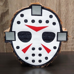 May include: A cardboard pinata shaped like a hockey mask with black and white details and red accents. The mask has a black eye hole and a red triangle on the forehead.