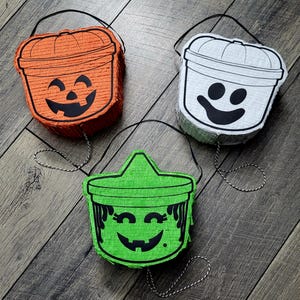 May include: Three Halloween-themed piñatas in orange, white, and green. Each piñata is bucket-shaped with a black face design and a black and white string handle. The orange piñata has a jack-o'-lantern face.