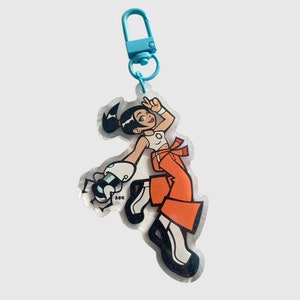 May include: A blue keychain with a cartoon character wearing an orange jumpsuit and holding a robotic arm. The character has black hair and is holding a peace sign.