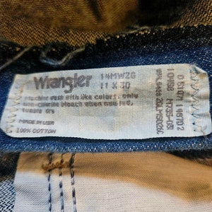 May include: A white Wrangler jeans care label with the text "Machine wash like colors, one wash-cycle, bleach, always wash cold, tumble dry, Made in USA, 100% Cotton".