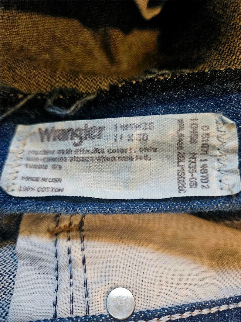 May include: A white Wrangler jeans care label with the text "Machine wash like colors, one wash-cycle, bleach, always wash cold, tumble dry, Made in USA, 100% Cotton".