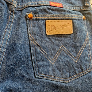 May include: Close-up of the back pocket of a pair of blue denim jeans. The pocket has a brown leather patch with the word "Wrangler" stitched in white. The pocket is stitched with orange thread.