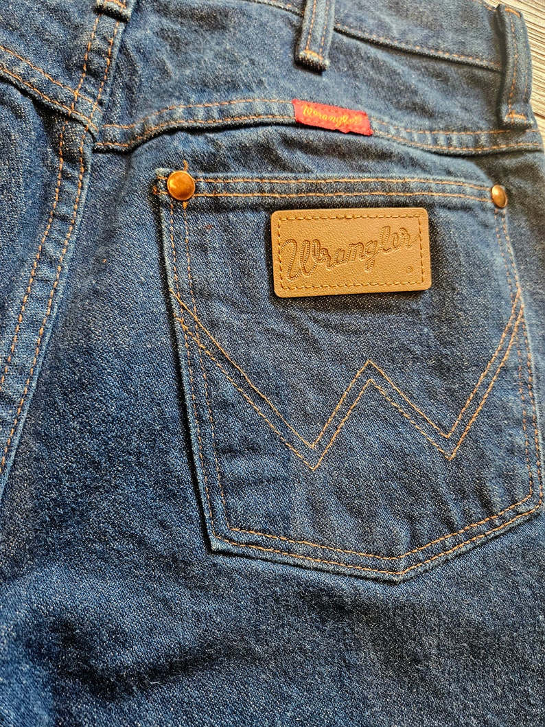 May include: Close-up of the back pocket of a pair of blue denim jeans. The pocket has a brown leather patch with the word "Wrangler" stitched in white. The pocket is stitched with orange thread.
