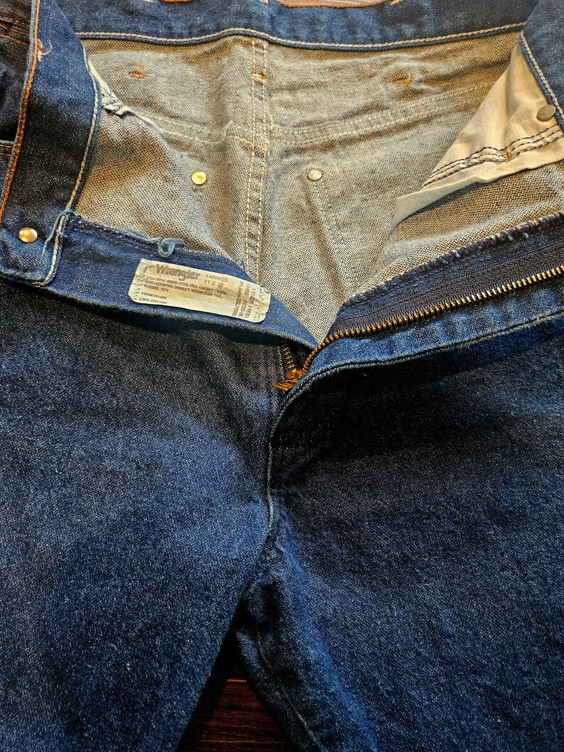 May include: A pair of blue denim jeans with a zipper fly and a Wrangler label. The jeans are unbuttoned and the waistband is folded down.