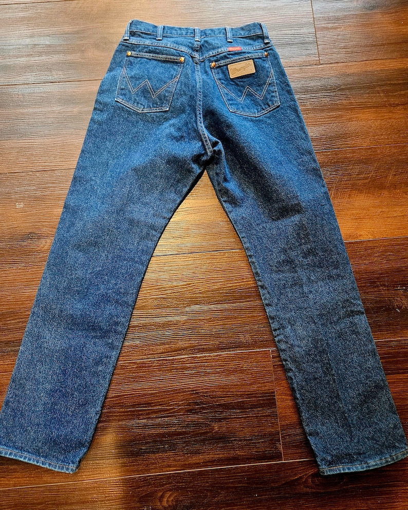 May include: A pair of blue denim jeans with a brown leather Wrangler label on the back pocket.