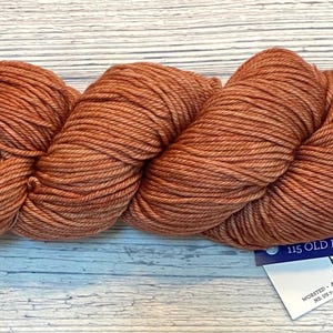 Peut inclure: Une pelote de laine couleur Vieux Rose, une teinte chaude et terreuse, est présentée sur une surface claire à grain de bois. La laine est étroitement enroulée, montrant la texture des fibres. Une étiquette avec le texte "115 OLD ROSE By Lisa Hannes" est attachée.