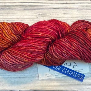 Peut inclure: Une pelote de laine rouge et orange vif, avec une étiquette indiquant "058 Zinnias". La laine est épaisse et texturée, idéale pour les projets de tricot ou de crochet. La laine mesure environ 10 cm de long.