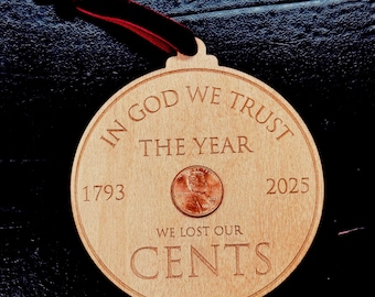 May include: Wooden ornament with the phrase "IN GOD WE TRUST" and "WE LOST OUR CENTS" engraved. A penny is set in the center. The years 1793 and 2025 are also engraved. A red ribbon is attached for hanging.
