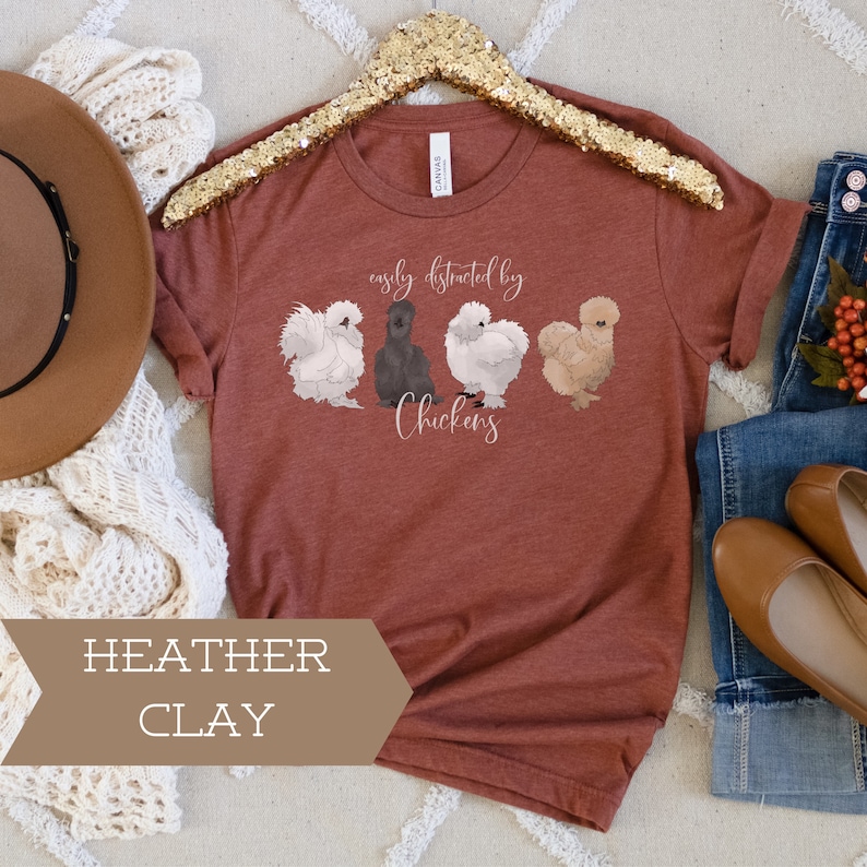 May include: A rust-colored t-shirt with the text "easily distracted by chickens" and a graphic of four chickens. The shirt is on a white surface with a brown hat and a white knitted scarf.