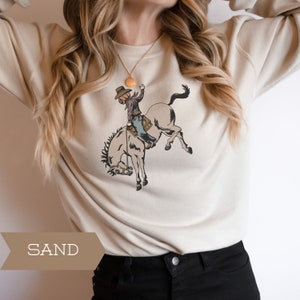 May include: Sand-colored sweatshirt featuring a graphic of a cowboy with a pumpkin head riding a bucking bronco. The cowboy is wearing a hat, shirt, and jeans. The word "SAND" is printed in a brown banner at the bottom.