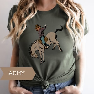 May include: Army green t-shirt featuring a cartoon illustration of a cowboy with a pumpkin head riding a bucking bronco. The cowboy is wearing a brown hat, purple shirt, blue jeans, and brown boots. The shirt has the word "ARMY" printed on it.