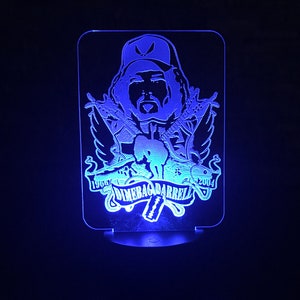 Puede incluir: Una luz nocturna LED azul con la silueta de Dimebag Darrell, un famoso guitarrista, con su guitarra característica y alas. El texto "1966 Dimebag Darrell 2004" está impreso debajo de la imagen.