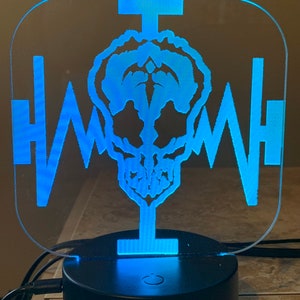 May include: A blue LED night light featuring a skull design with a heartbeat-like pattern. The light is mounted on a black circular base with a power cord.