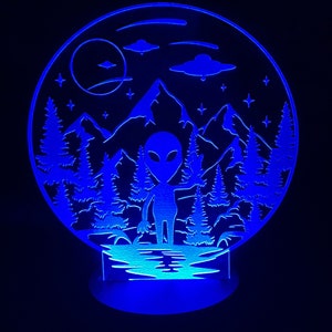 May include: Blue LED night light with a silhouette of an alien standing in a forest with mountains and UFOs in the background.