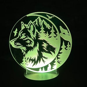 May include: A glowing, circular LED night light featuring a wolf howling at the moon. The design includes mountains, trees, and birds, all etched into a clear acrylic panel. The base is a small, round metal stand.