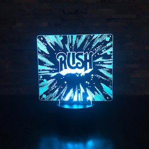 Può includere: Una luce notturna LED blu con una base in acrilico trasparente. La luce presenta una grafica stilizzata della parola "RUSH" con un effetto a stella.