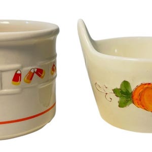 May include: A cream-colored ceramic crock featuring a horizontal band of candy corn motifs and an orange stripe at the base, alongside a cream-colored ceramic bowl with two handles and a painted orange pumpkin with green leaves on the front.
