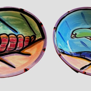 May include: Two ceramic bowls with hand-painted designs. One bowl features a pink and red seashell on a light blue and green background. The other bowl features a green dolphin swimming in blue water.
