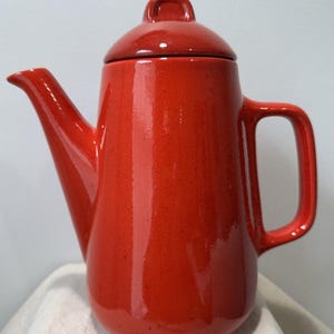 May include: A bright red ceramic teapot with a glossy finish, featuring a tall cylindrical body, a curved spout, a sturdy side handle, and a matching removable lid with a small knob handle on top, showcasing a smooth, solid-colored surface.