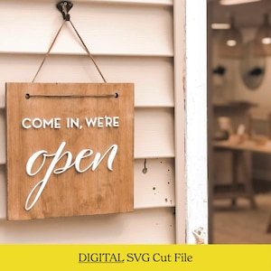 以下が含まれることがあります： 白い文字で「Come in, we're open」と書かれた木製の看板。白い壁のフックに掛けられています。