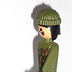 May include: A green camouflage plastic toy soldier with a black machine gun and a green knit hat. The soldier is wearing black boots.
