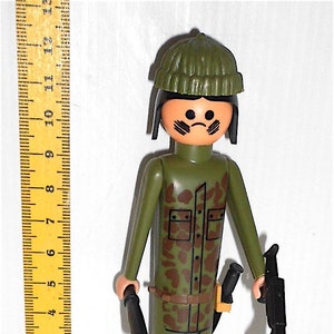 May include: A green plastic toy soldier figure wearing a green camouflage uniform, a black hat, and holding a black rifle and a black baton.