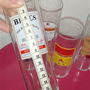 May include: Four clear glass tumblers with labels for different brands of whisky. The tumblers are on a red surface and are being measured with a tape measure. The tape measure reads 18 inches.