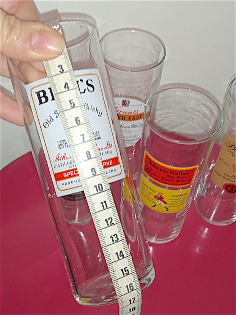 May include: Four clear glass tumblers with labels for different brands of whisky. The tumblers are on a red surface and are being measured with a tape measure. The tape measure reads 18 inches.