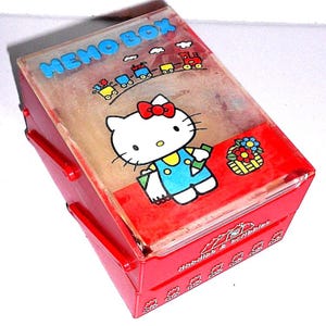 Puede incluir: Caja de notas de plástico roja con una tapa transparente que presenta una ilustración de Hello Kitty. La tapa tiene un diseño de tren y el texto "MEMO BOX". La caja tiene un cajón con el texto "Huddles & Scribbles".