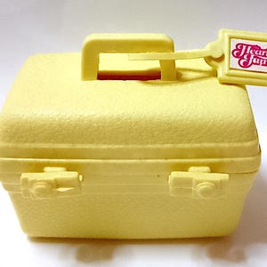 May include: A small, yellow, vintage toy suitcase with a textured surface. It has a handle, two clasps, and a tag that reads "Heart Family" in pink and white. The suitcase is approximately 10 cm wide.