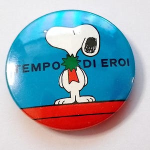 Può includere: Un bottone rotondo blu e rosso con un'immagine di cartone animato di Snoopy che indossa una ghirlanda verde e un nastro rosso. Il testo "TEMPO DI EROI" è stampato in nero sul bottone.
