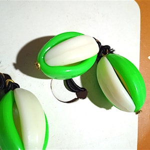 Puede incluir: Primer plano de tres pares de pendientes. Cada pendiente presenta una cubierta exterior verde brillante, de forma ovalada, con una pieza interior oblonga blanca. Los pendientes están conectados por cordones negros y pequeños detalles dorados. Sobre una superficie blanca.