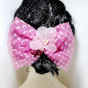 Può includere: Un fiocco rosa per capelli con pois bianchi e un fiore come ornamento. Il fiocco è realizzato in tessuto trasparente ed è attaccato a un'acconciatura nera. Il fiore è di una tonalità di rosa più chiara con glitter.