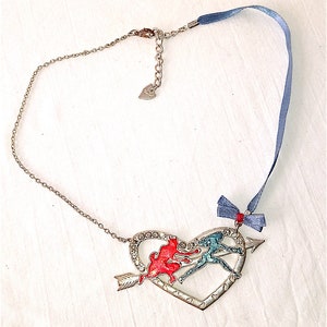 Op de afbeelding: Een zilveren hartvormige ketting met een rood en blauw emaille ontwerp van twee figuren die een pijl vasthouden. De ketting heeft een zilveren ketting en een blauw lint met een strik.