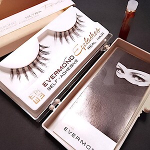 Può includere: Primo piano di un set di ciglia autoadesive Evermond in una scatola bianca e beige. La scatola è aperta, rivelando le ciglia e un piccolo flacone. Il testo "EVERMOND Eyelashes REAL HAIR" è visibile.
